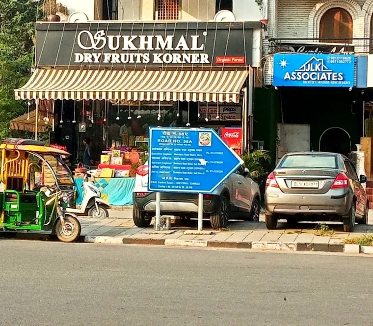 The shop seen from across the road, its striped awning and navy signboard behind parked cars and an e-rickshaw